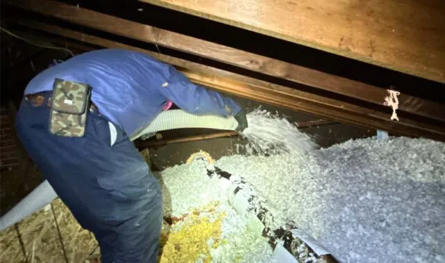 A DeVere Insulation technician wearing a blue uniform and gloves uses a large flexible hose to blow loose-fill fiberglass insulation into a dark residential attic.