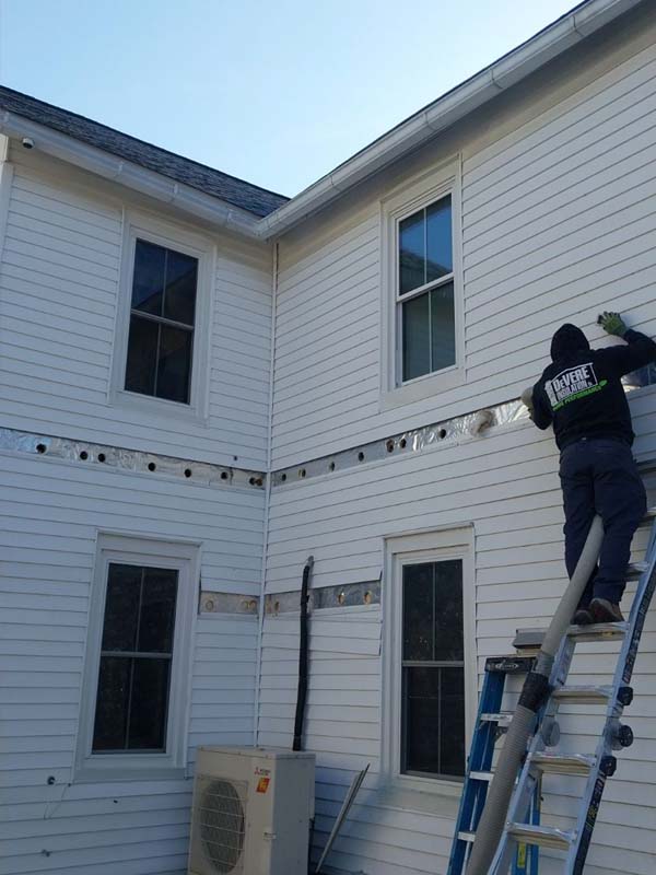 DeVere Insulation technician injecting blown-in cellulose insulation into a home’s upper wall using a hose from a ladder as part of a drill-and-fill retrofit.