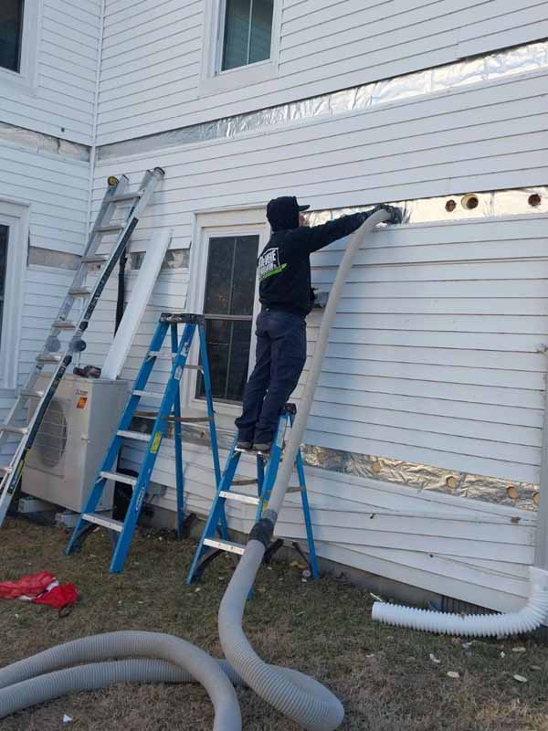 DeVere Insulation technician performing drill-and-fill insulation on a home’s exterior wall while standing on a step ladder, surrounded by tools and insulation hoses.