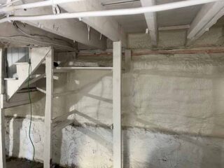 Spray foam insulated basement wall beneath wooden stairs and joists.