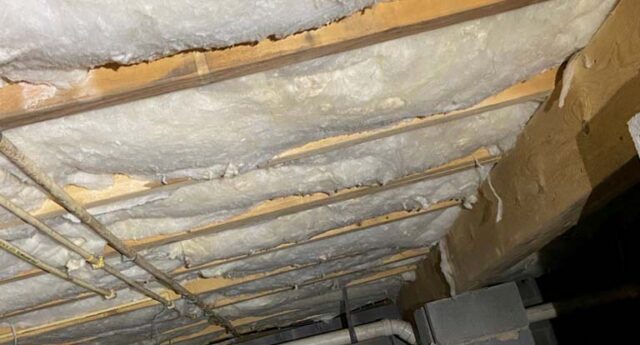 Spray foam insulation applied to crawl space ceiling between wooden joists with exposed plumbing pipes underneath.