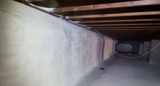 Spray foam insulation applied to crawl space walls and ceiling, with exposed pipes and vapor barrier on the floor.