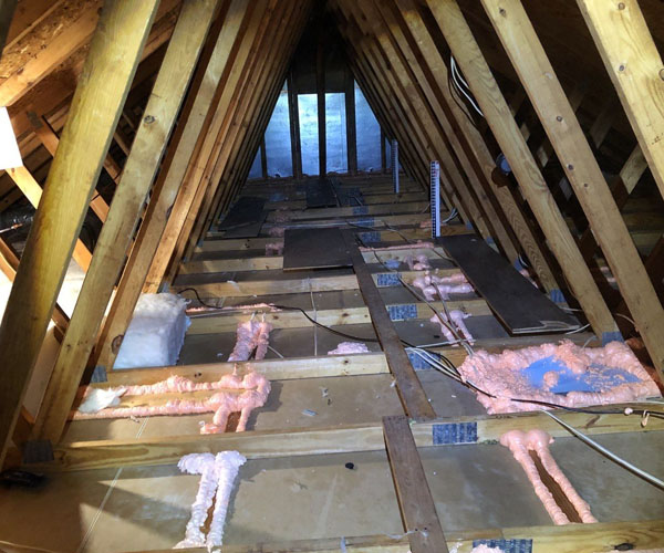 n interior view of an unfinished attic showing pink expanding spray foam used to seal gaps and penetrations around wooden floor joists before adding insulation.