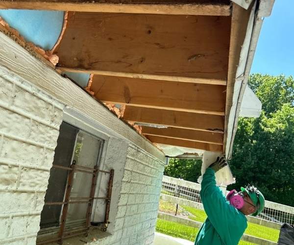 A construction worker wearing a respirator mask and hard hat installs fiberglass batt insulation into the open wooden joists of a cantilevered soffit overhang.